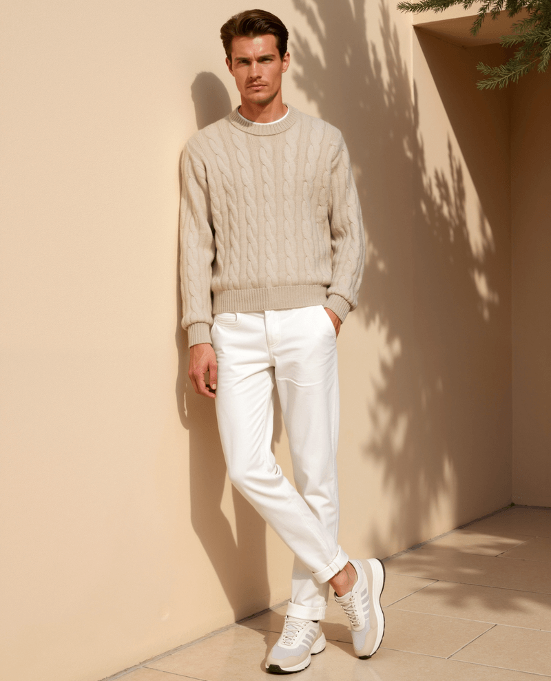 Man wearing a beige sweater and white pants standing against a beige wall with a plant in the corner.