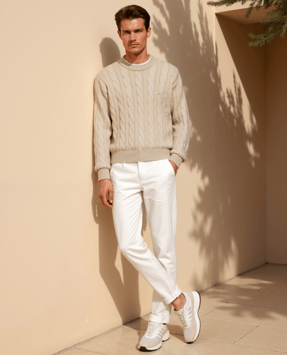 Man wearing a beige sweater and white pants standing against a beige wall with a plant in the corner.