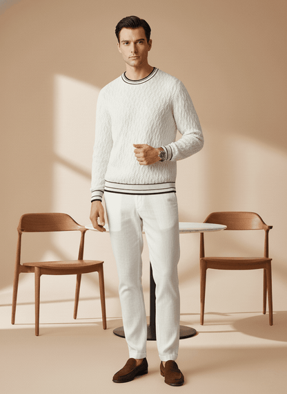 Man wearing a white sweater and pants in a minimalistic room with wooden chairs and a table.