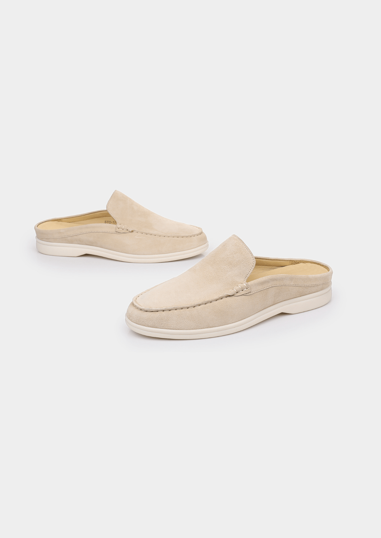 A backless slip-on loafer made of light beige sheep suede with hand-stitched detailing and a cream-colored rubber sole.
