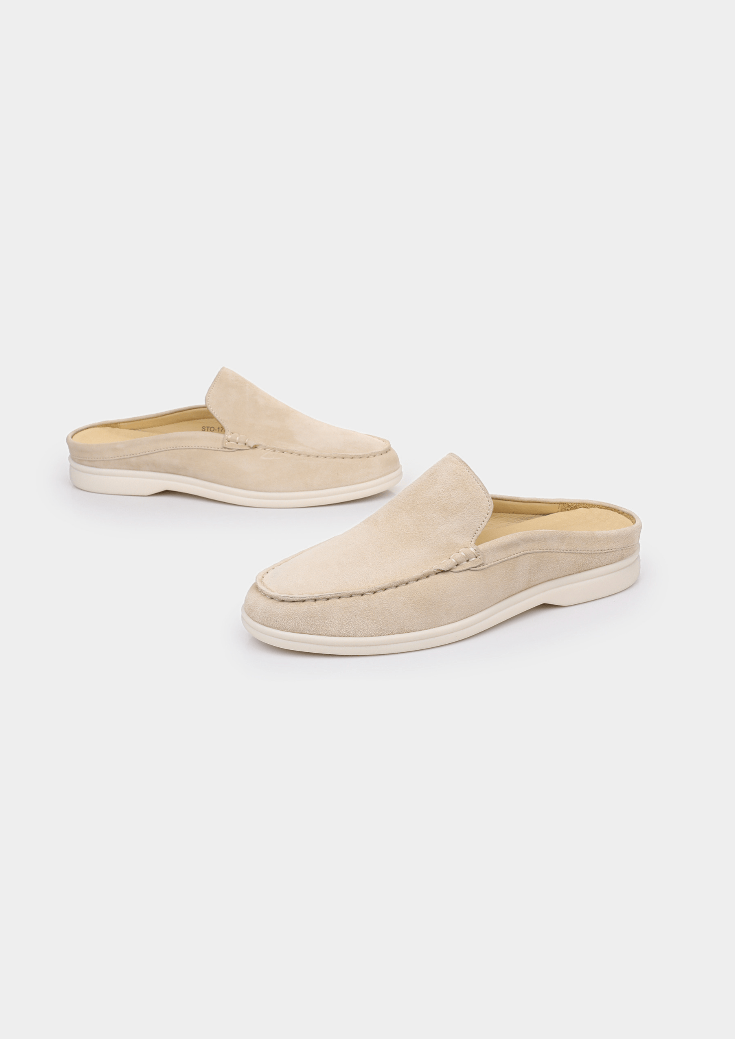 A backless slip-on loafer made of light beige sheep suede with hand-stitched detailing and a cream-colored rubber sole.