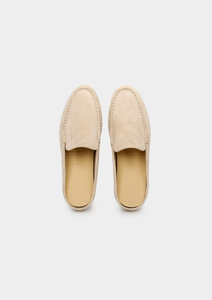 A backless slip-on loafer made of light beige sheep suede with hand-stitched detailing and a cream-colored rubber sole.