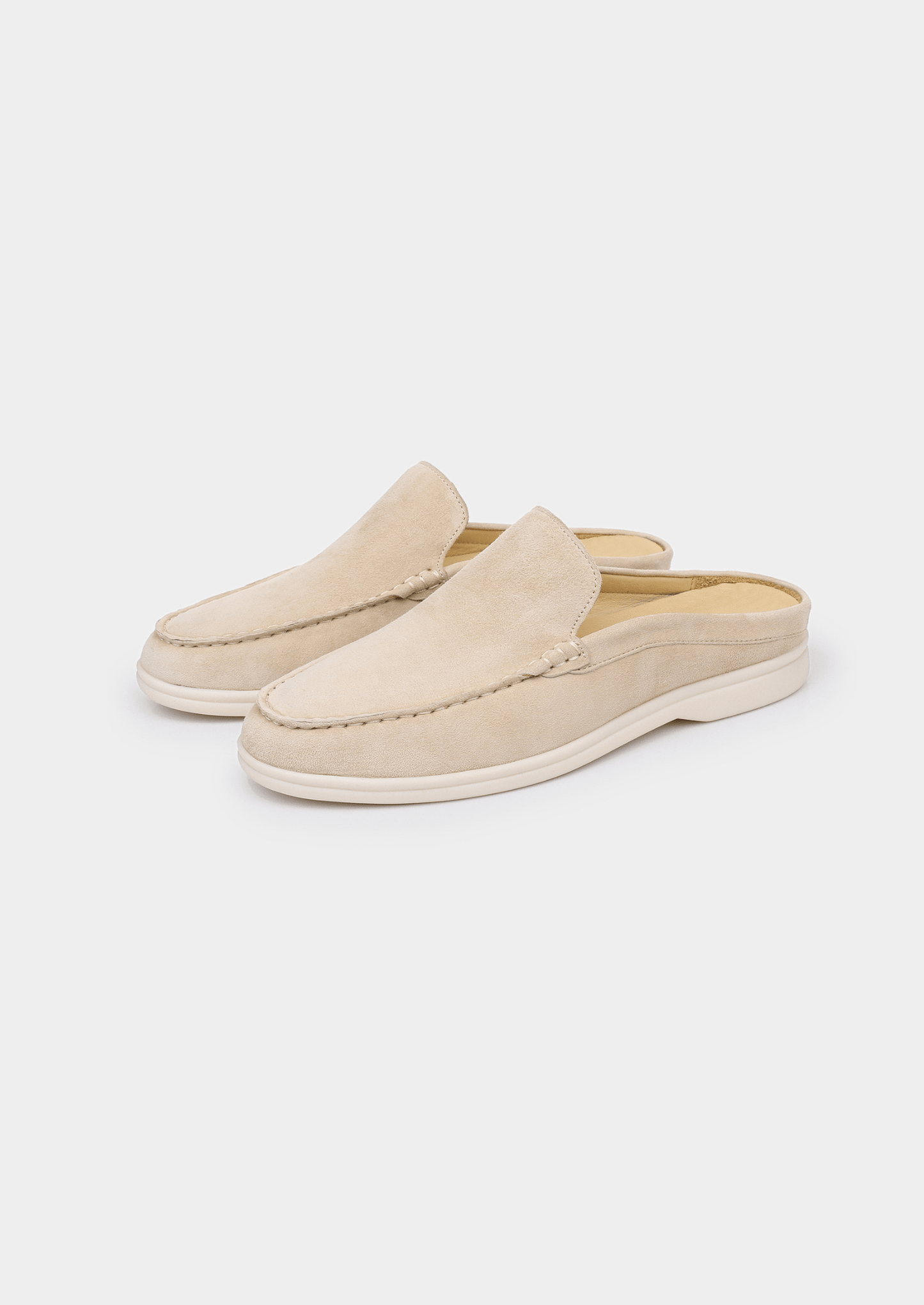 A backless slip-on loafer made of light beige sheep suede with hand-stitched detailing and a cream-colored rubber sole.