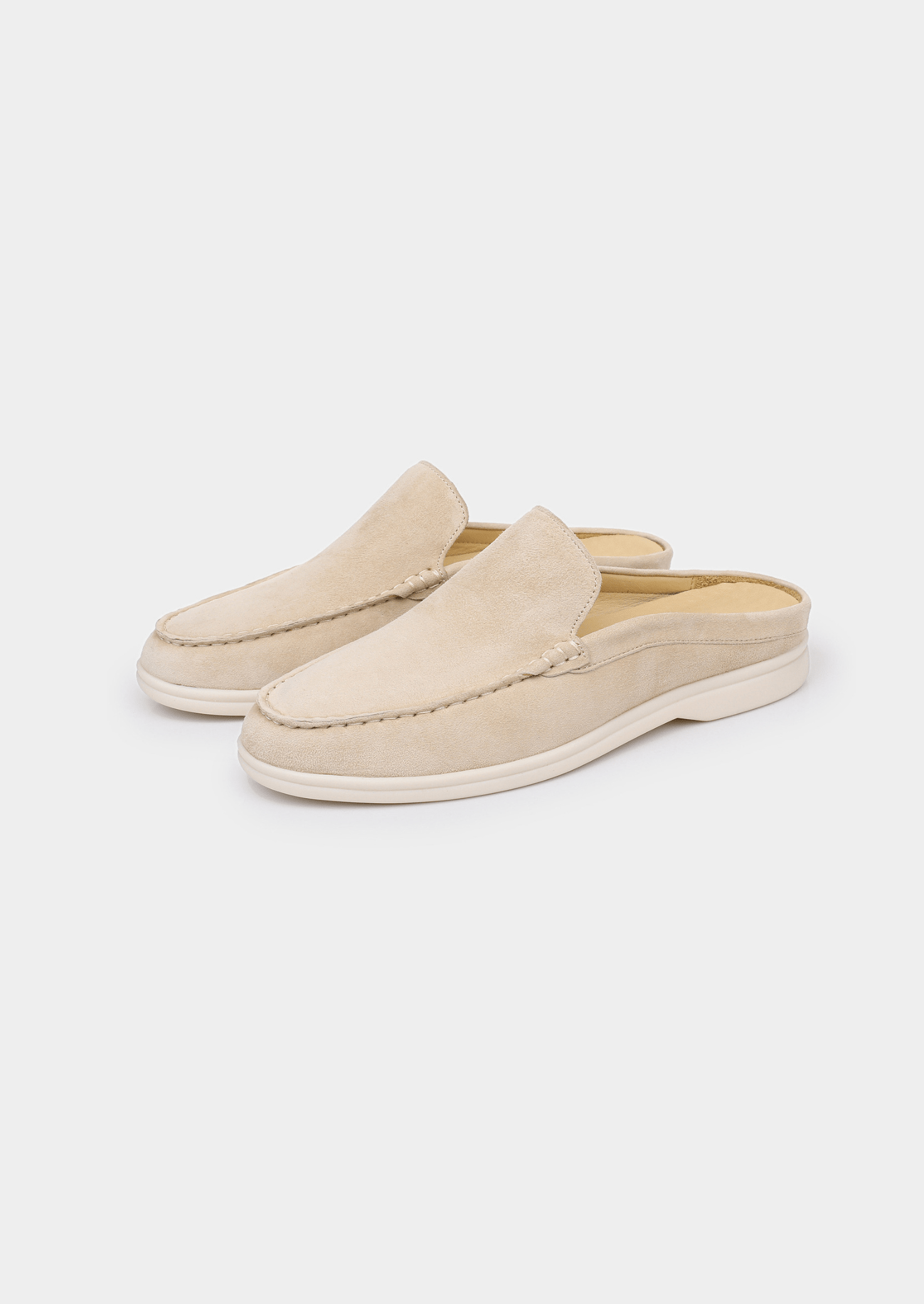 A backless slip-on loafer made of light beige sheep suede with hand-stitched detailing and a cream-colored rubber sole.