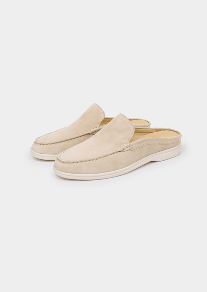 A backless slip-on loafer made of light beige sheep suede with hand-stitched detailing and a cream-colored rubber sole.