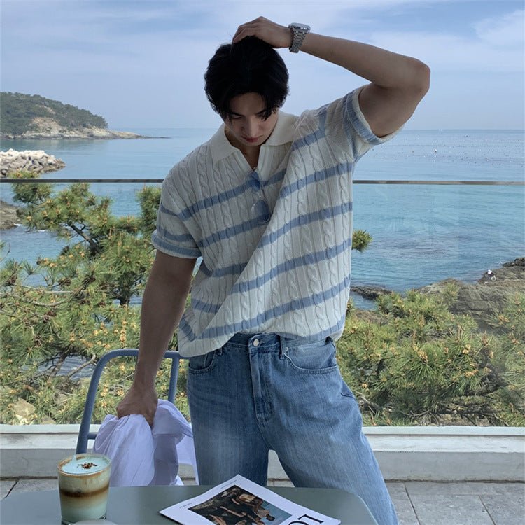 A man wearing a relaxed fit, short-sleeved cable knit polo in cream with blue stripes, sitting at an outdoor table overlooking the ocean. The shirt features a modern boxy silhouette and a dropped shoulder design.
