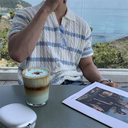 A man wearing a relaxed fit, short-sleeved cable knit polo in cream with blue stripes, sitting at an outdoor table overlooking the ocean. The shirt features a modern boxy silhouette and a dropped shoulder design.